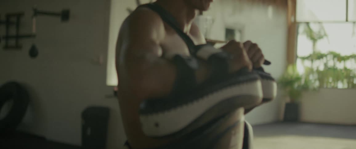 Close up side view of a muscular shirtless male trainer with kick pads and a body protector in an outdoor gym
