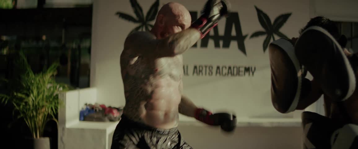 A muscular, tattooed fighter practices powerful Muay Thai strikes on pads held by his coach in cinematic slow motion