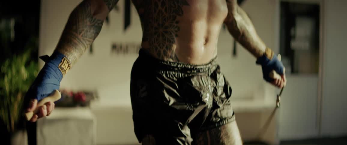An intense slow-motion close-up focuses on the defined abs of a shirtless, tattooed boxer as he skips rope in a gym