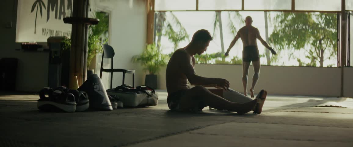 A male fighter puts on his shin guards while his training partner jumps rope in the background of a sunlit tropical gym