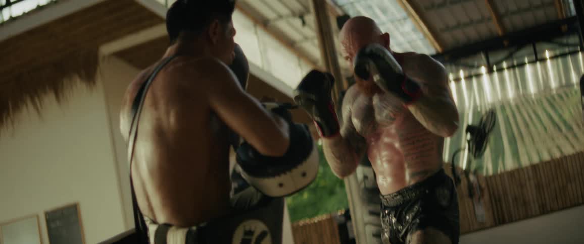 A tattooed Muay Thai fighter spars with his trainer in an intense, sweaty, slow-motion training session at a gym