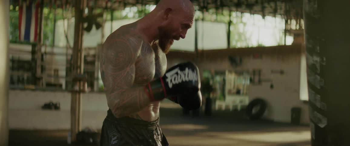 A powerful, heavily tattooed Muay Thai fighter intensely trains with punches and kicks on a heavy bag in a gritty, open-air gym in slow motion