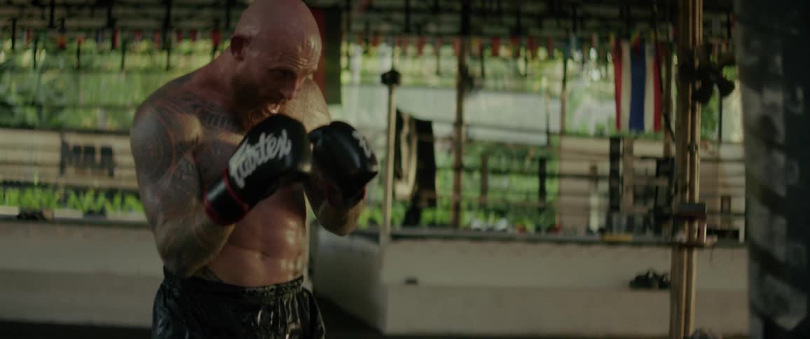 A heavily tattooed and muscular fighter relentlessly punches and kicks a heavy bag during an intense slow-motion training session in a gym