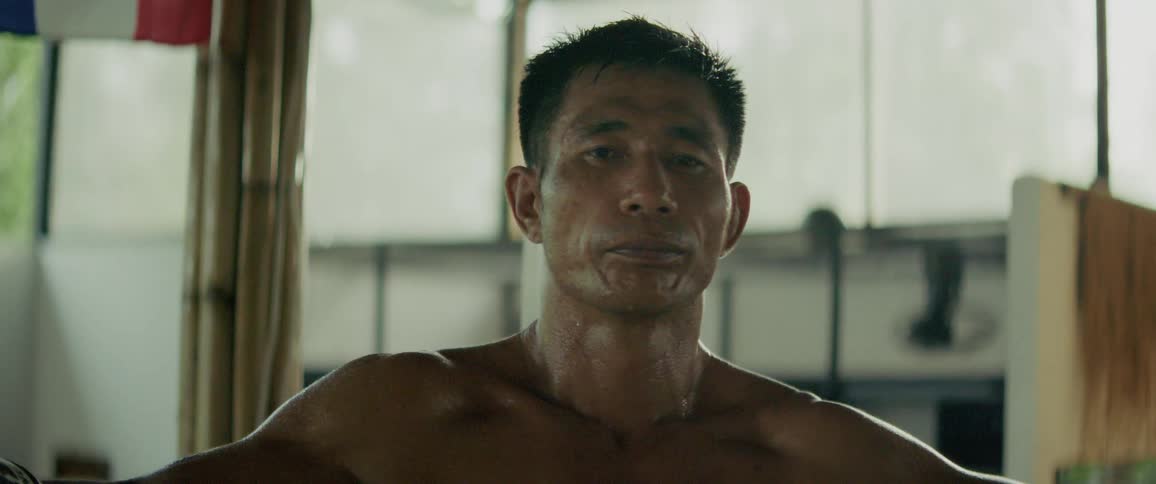 A close-up shot of a determined, sweaty, shirtless man staring directly into the camera before breaking into a slight, satisfied smile