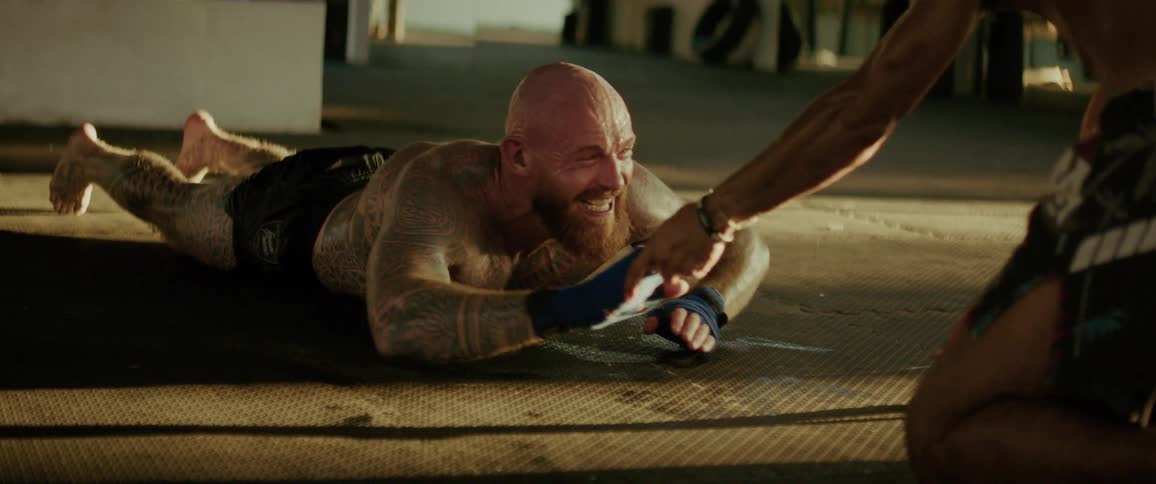 A heavily tattooed fighter collapses from exhaustion but, with his trainer's encouragement, pushes through to finish his workout, ending with a smile and a respectful hug