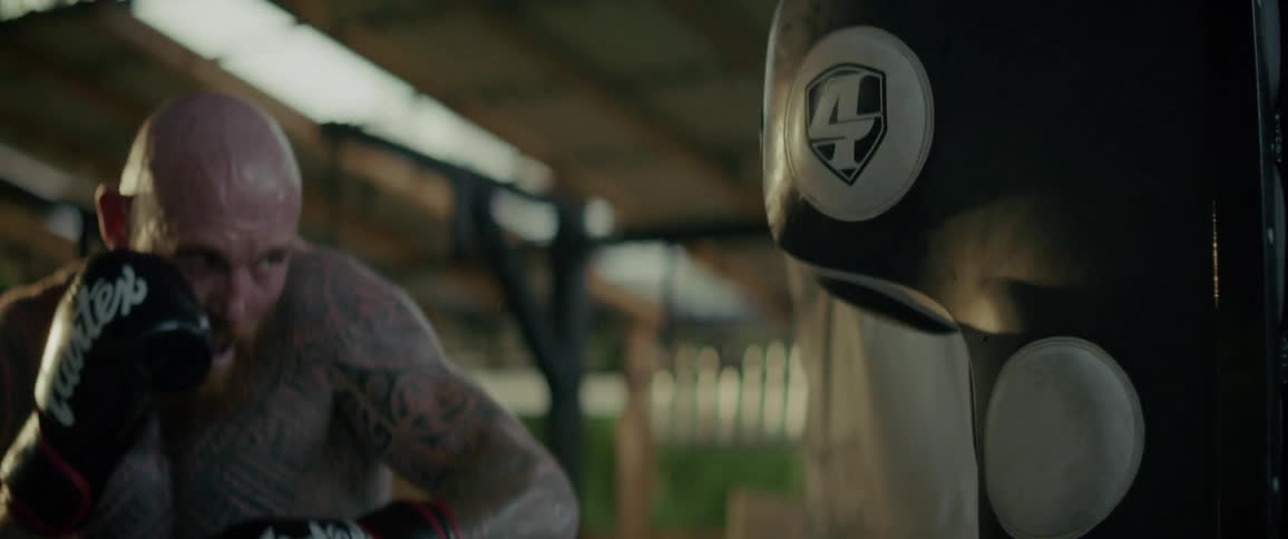 A powerful, tattooed man with a red beard intensely trains in slow motion, landing heavy punches on a standing bag at an outdoor gym