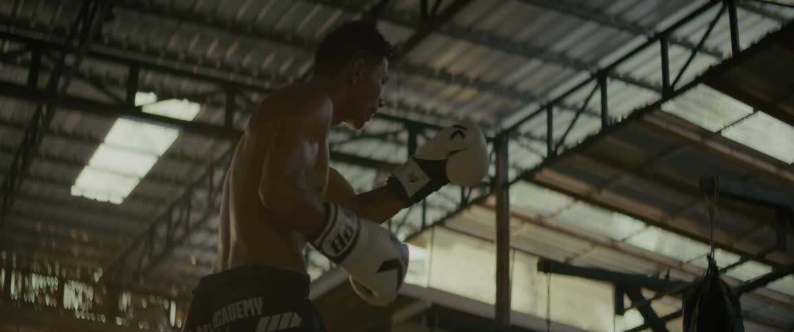 A muscular Muay Thai fighter practices powerful kicks and shadowboxing in a sun-drenched gym, captured in dramatic slow motion