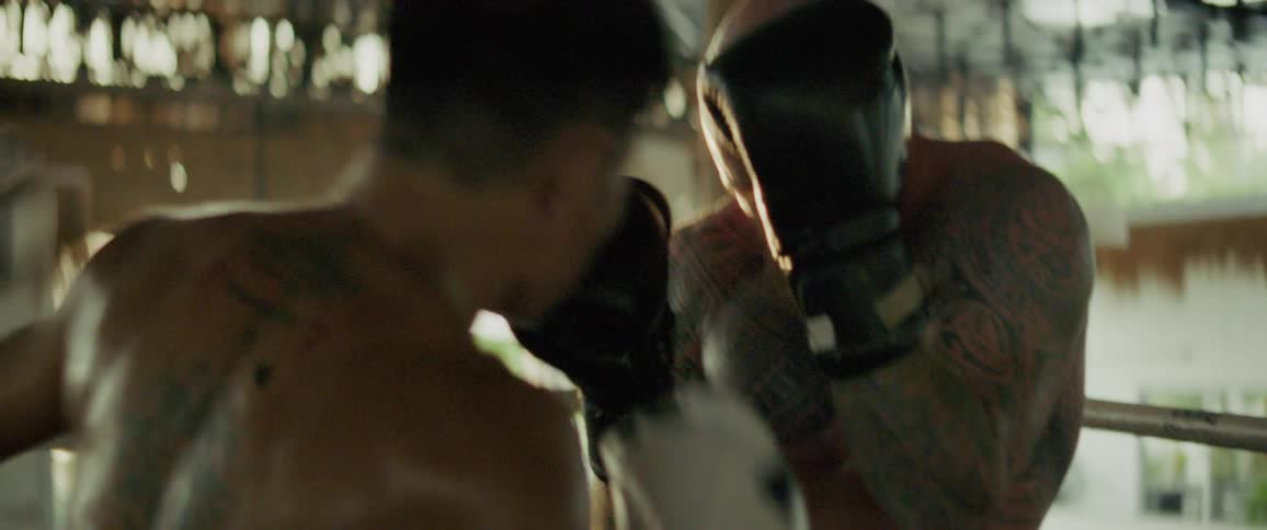 A heavily tattooed boxer with a red beard spars intensely in the ring, blocking and taking punches in dramatic slow motion