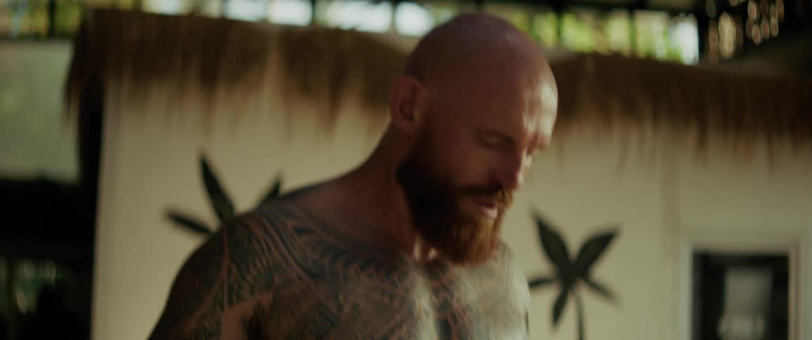 A heavily tattooed and muscular bald man with a red beard intensely jumps rope during a workout in a tropical outdoor gym in slow motion