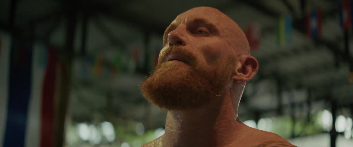 A slow-motion close-up pans up from the chiseled abs to the determined face of a sweaty, muscular, tattooed fighter in a gym