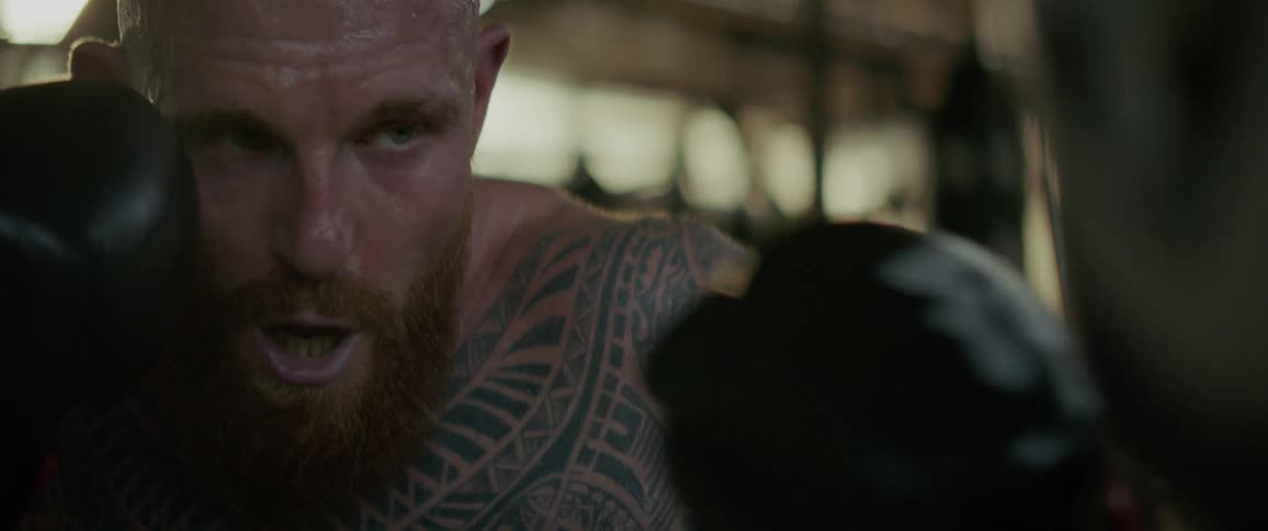 An intense, sweaty, tattooed boxer with a red beard trains with a punching bag in a gritty gym in slow motion