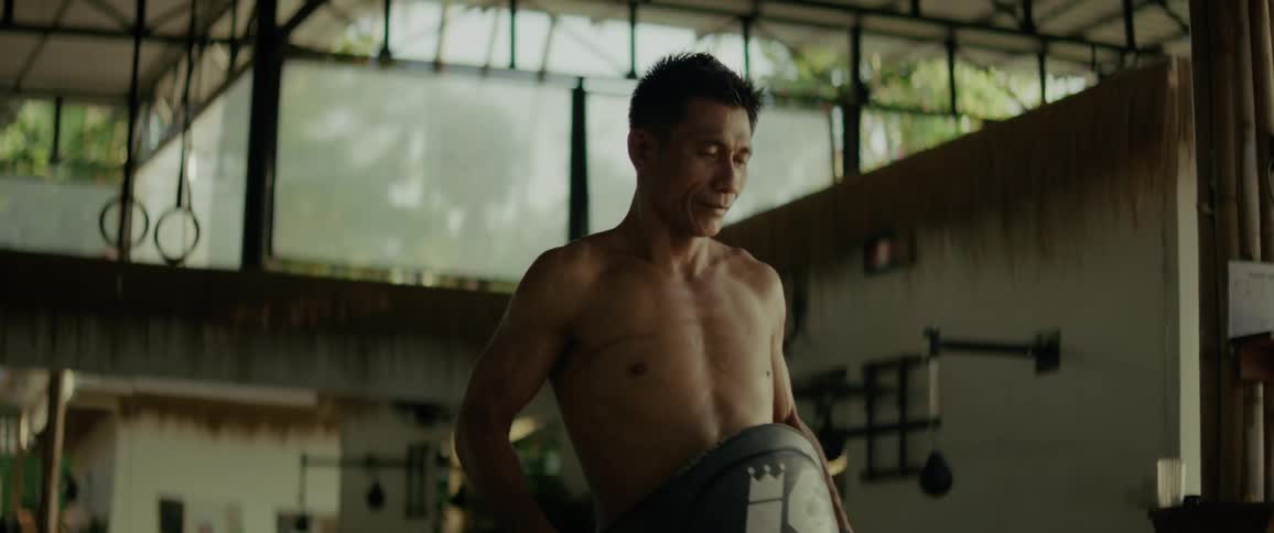 A muscular Asian Muay Thai trainer puts on a protective belly pad and picks up a large kicking shield in slow motion