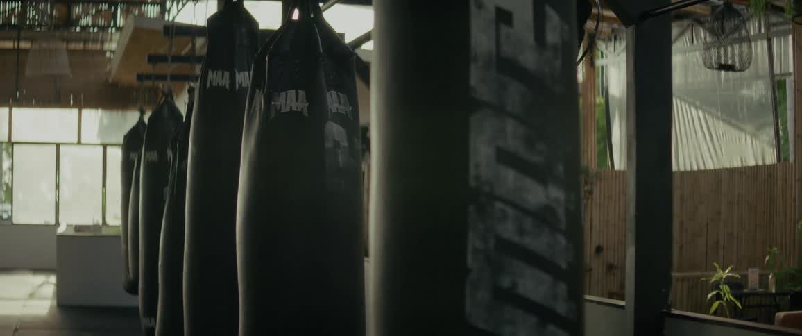 A row of black punching bags hangs silently in a dimly lit, empty boxing gym