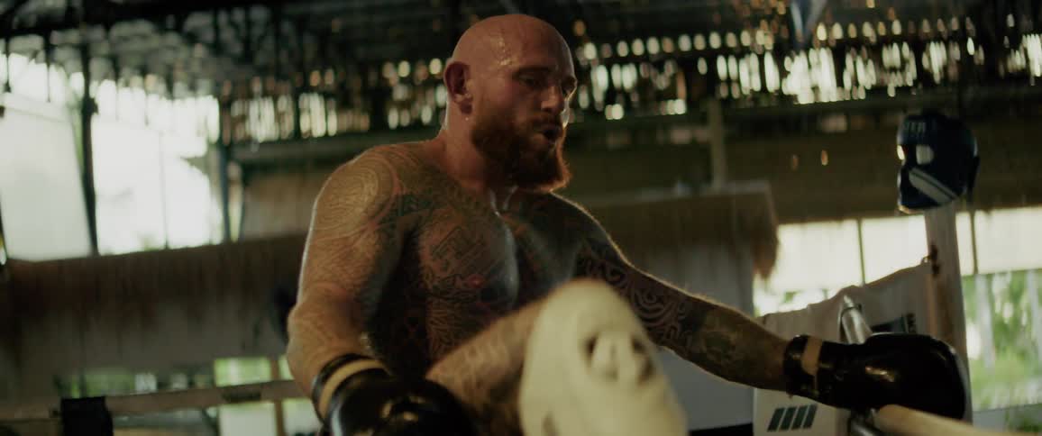 A heavily tattooed and muscular fighter with a red beard stretches intensely in a rustic boxing ring in slow motion