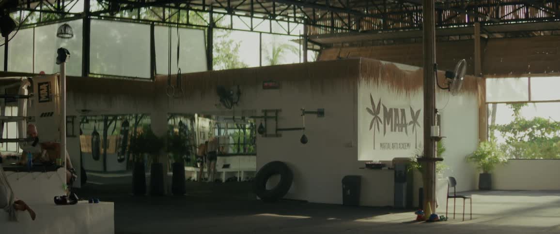 A wide, static shot captures the interior of a sunlit, tropical-style Martial Arts Academy with a lone athlete training in the background