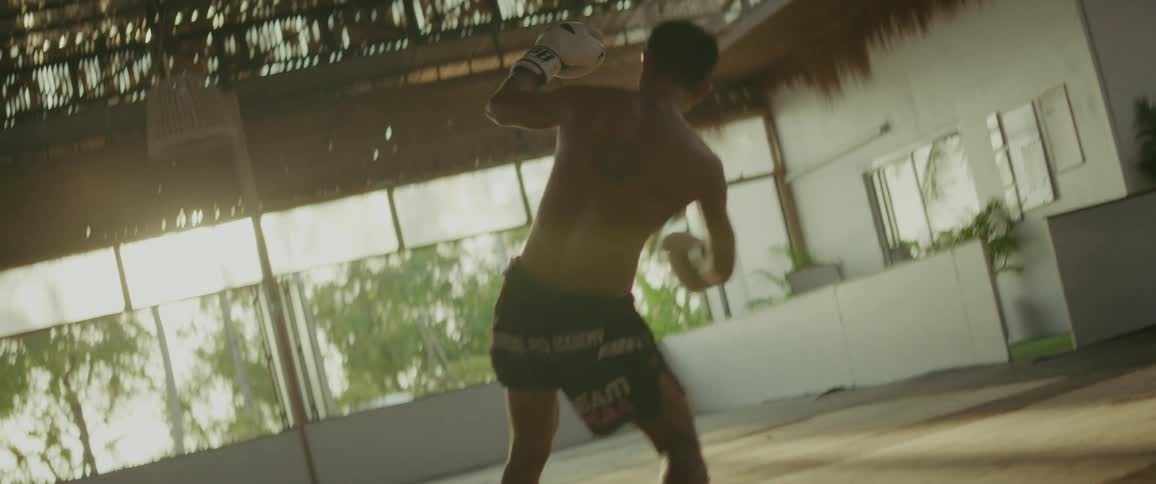 A determined martial artist practices powerful kicks and strikes in a sun-drenched gym, captured in dramatic slow motion