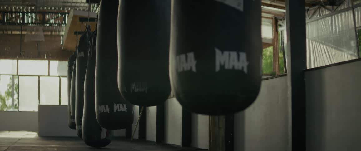 A moody, slow-motion tracking shot reveals a row of dark punching bags hanging in an empty martial arts gym
