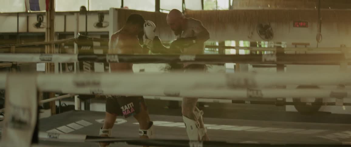 Two muscular Muay Thai fighters spar in a boxing ring as the bright sun flares through the gym windows in slow motion