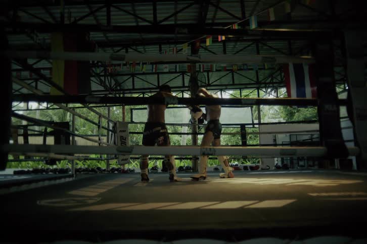 Two dedicated Muay Thai fighters train together, sparring in slow motion inside an atmospheric open-air boxing ring