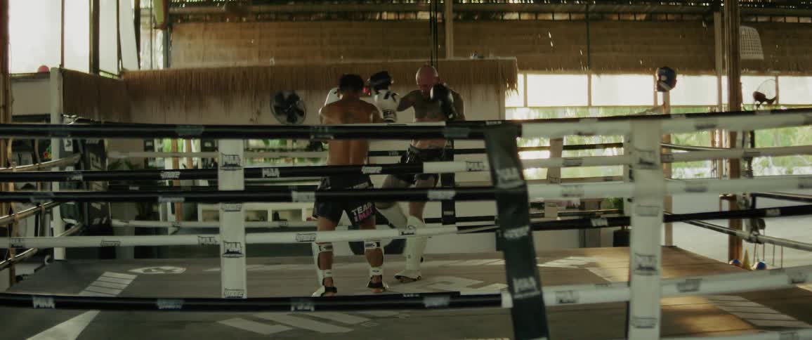 A young Muay Thai fighter knocks down his larger, tattooed opponent during an intense sparring session in a boxing ring