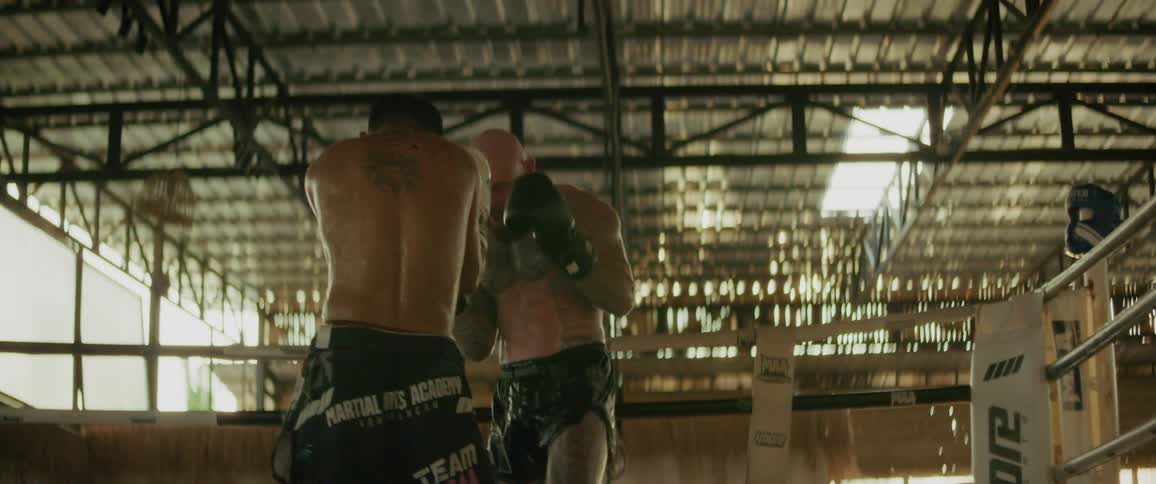 Two muscular male fighters engage in an intense sparring session in a gritty boxing ring, showcasing their kickboxing skills in slow motion