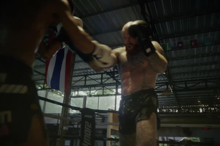 Intense slow-motion footage shows two muscular men engaged in a fierce Muay Thai sparring session in a rustic boxing ring