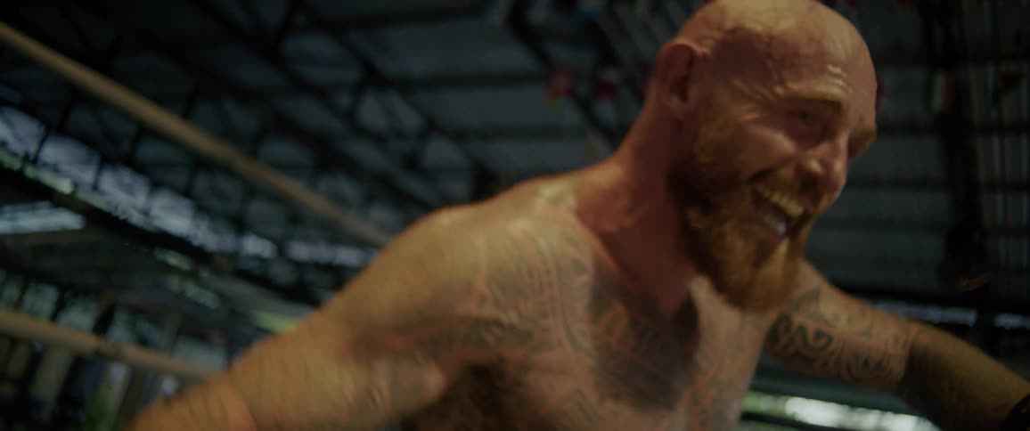A muscular, tattooed boxer rises from the mat in slow motion, grimacing with extreme exertion and determination during an intense training session