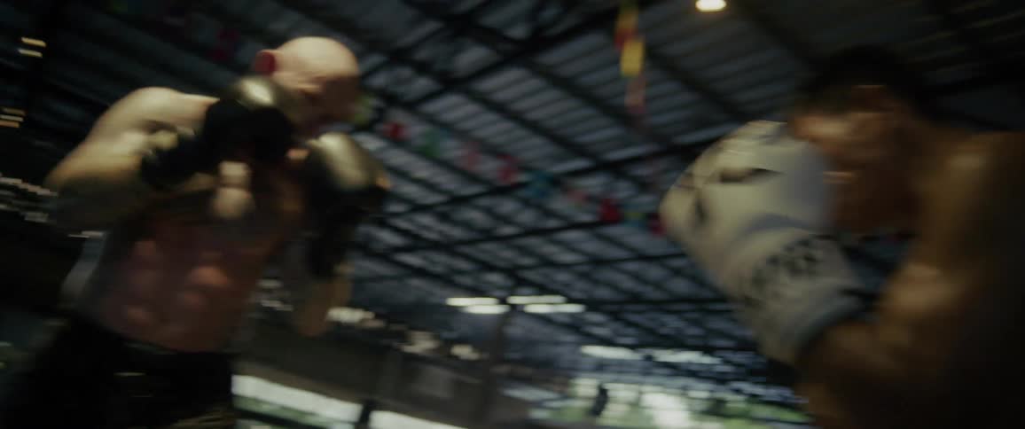 A muscular tattooed boxer and his opponent spar intensely in a slow-motion training session inside a dimly lit boxing ring