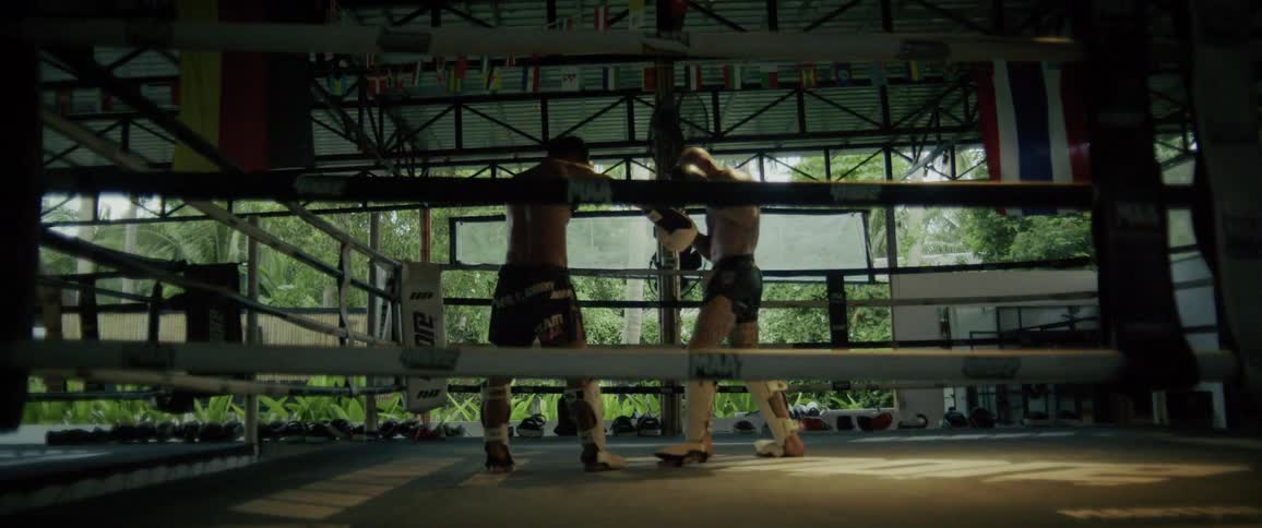 Two Muay Thai fighters spar in slow motion inside a boxing ring at a tropical outdoor gym