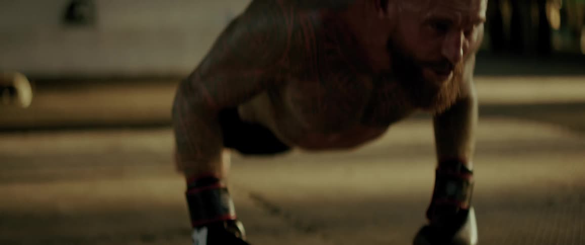 A heavily tattooed and muscular fighter with a beard intensely performs explosive push-ups with boxing gloves on in slow motion