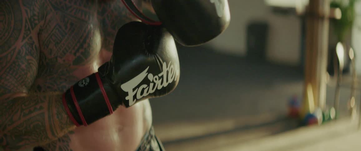 A muscular, tattooed fighter with a red beard tightens his boxing gloves with his teeth in a gritty, slow-motion shot