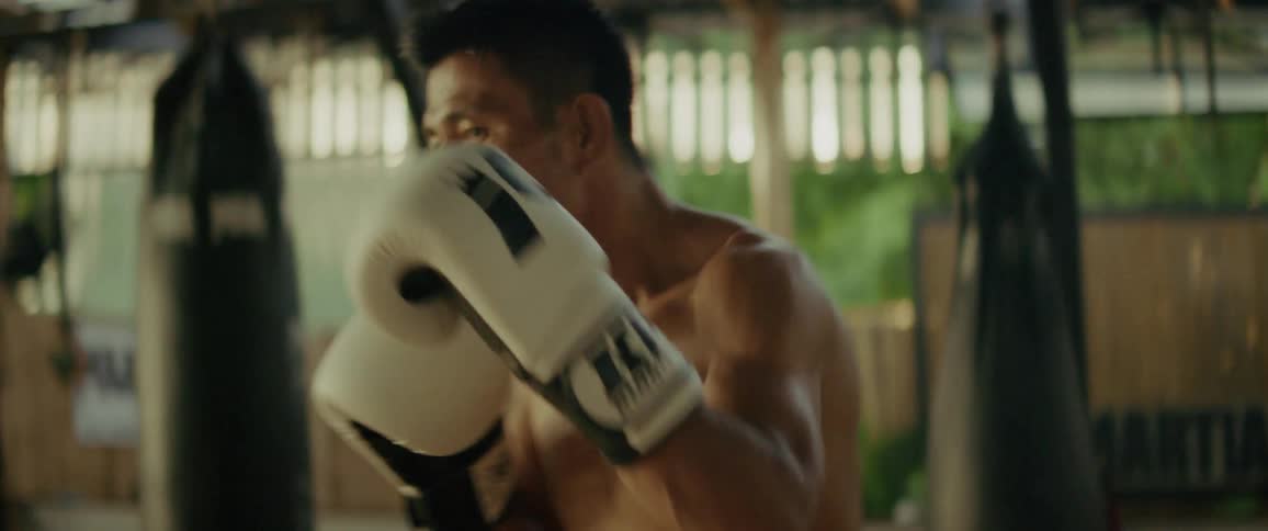 A focused, shirtless boxer trains intensely, throwing powerful punches in a gritty gym during a slow-motion sequence