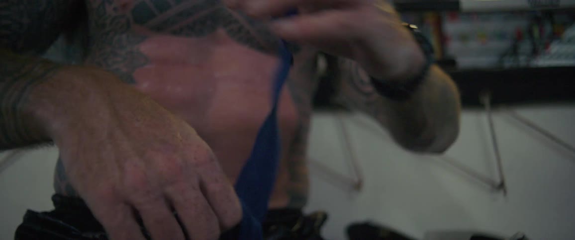 A heavily tattooed and muscular fighter with a red beard carefully wraps his hands with blue tape in slow motion before a training session