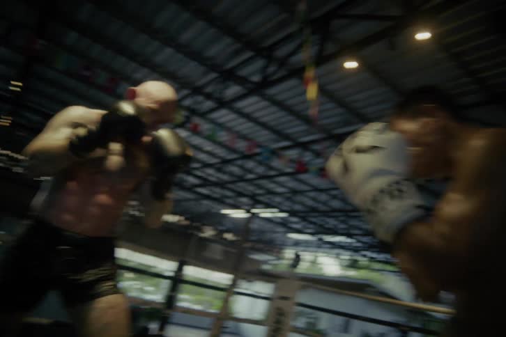 Two muscular boxers, one heavily tattooed with a red beard and the other his agile opponent, spar intensely in a dramatic slow-motion fight inside the ring