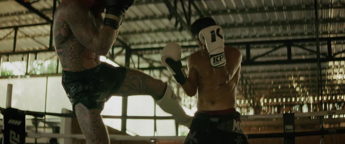 A heavily tattooed fighter and his opponent trade powerful kicks and punches during a slow-motion sparring match in a boxing ring