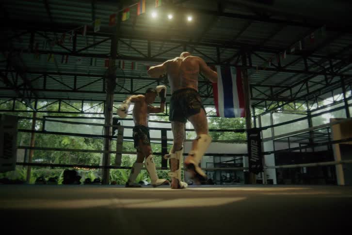 Slow motion footage shows two skilled Muay Thai fighters sparring intensely inside a dramatically lit boxing ring