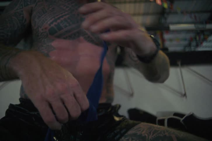 A heavily tattooed fighter with a red beard meticulously wraps his hands in a gritty boxing gym in slow motion