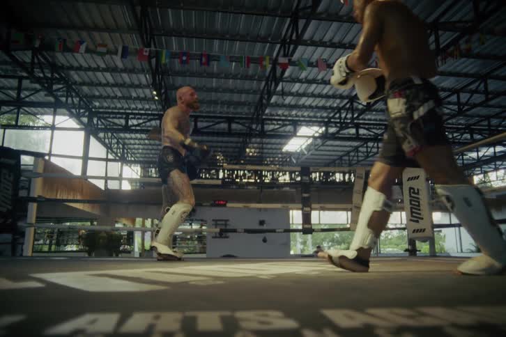 Two muscular Muay Thai fighters spar in a gritty boxing ring, ending in a dramatic slow-motion takedown