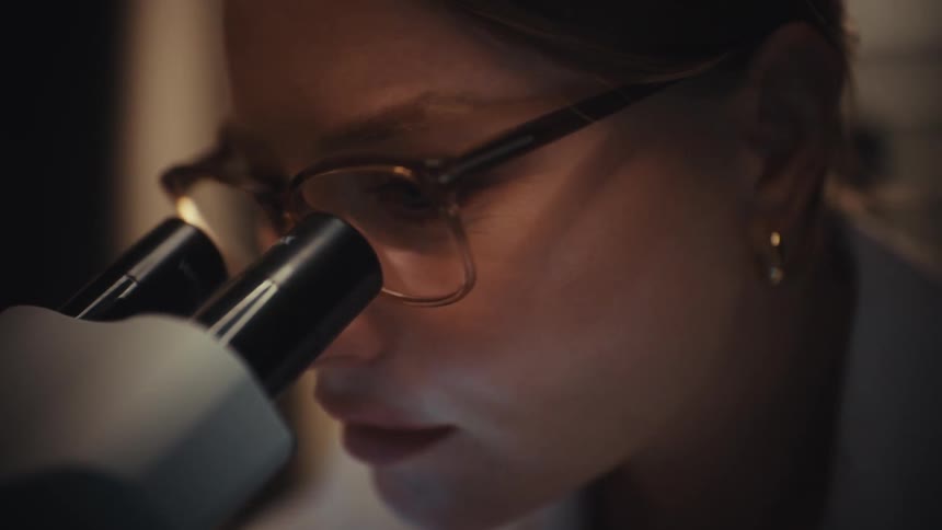 An intense close-up shows a focused female scientist looking through a microscope in a dark laboratory setting