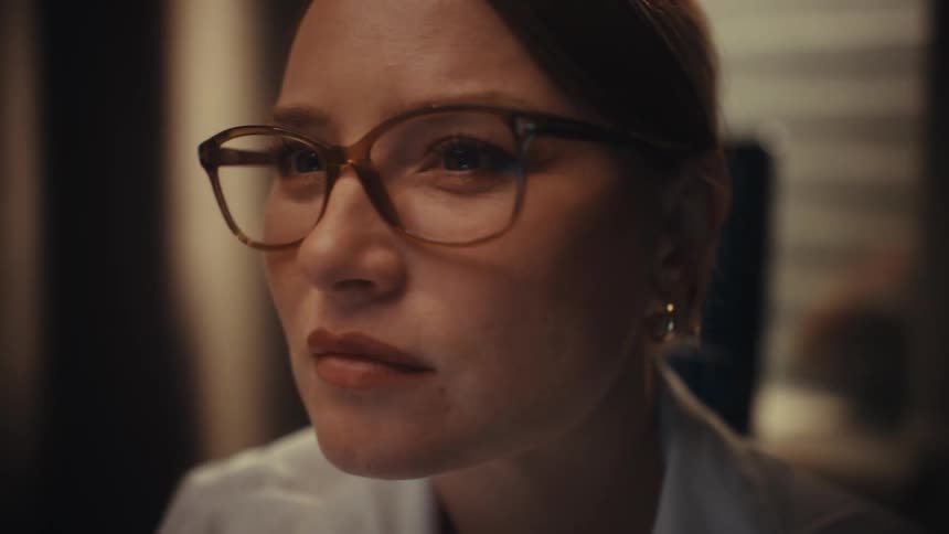An intense close-up of a serious woman with glasses looking intently at something off-screen in a dimly lit room