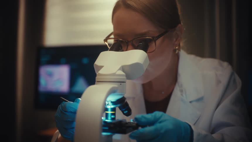 A focused female scientist meticulously examines a sample under a microscope in a dimly lit, modern laboratory