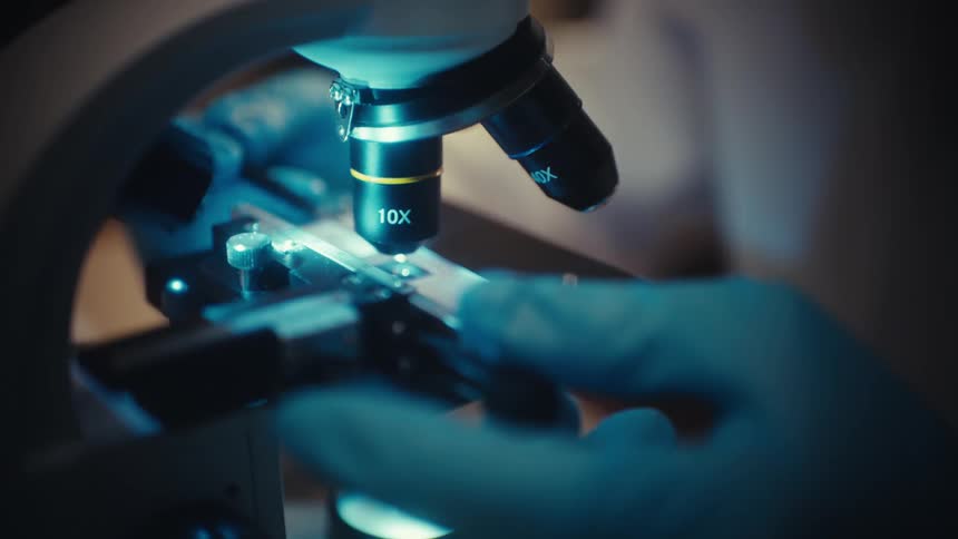 A close-up view of a scientist's gloved hands adjusting a specimen slide under a microscope in a lab