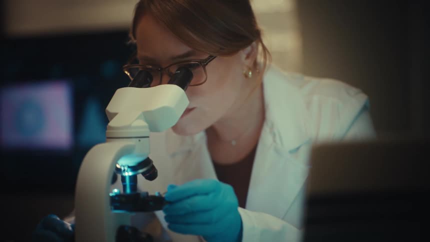 Female scientist in a white coat and glasses meticulously studies samples under a microscope in a modern research laboratory