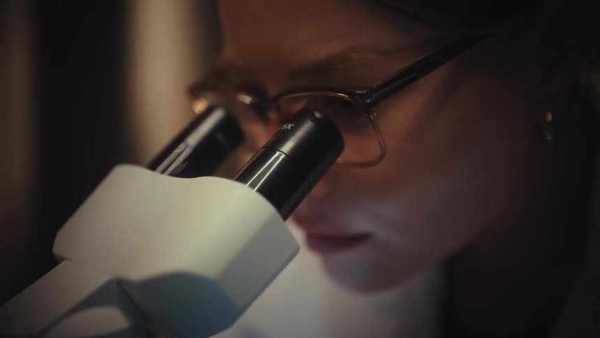A focused female scientist in glasses carefully examines a specimen slide under a modern microscope in a dimly lit laboratory