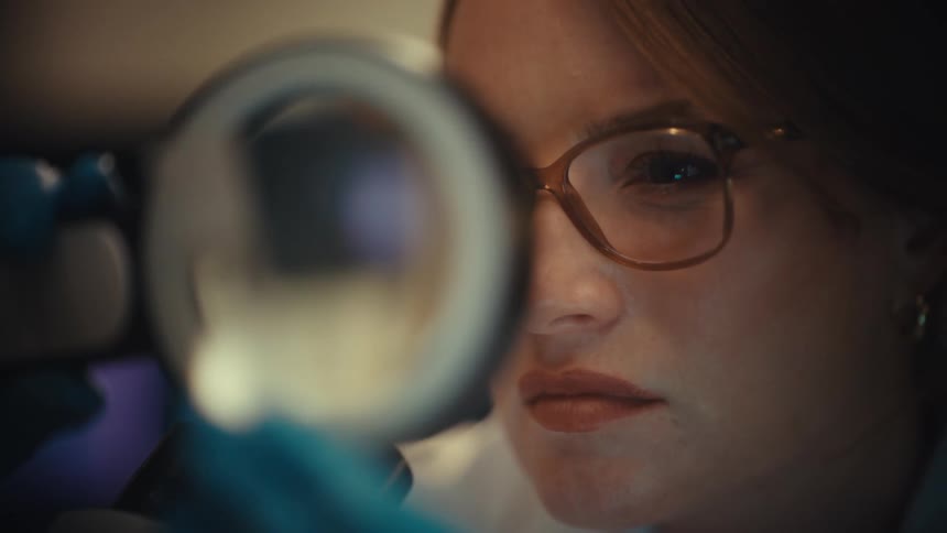 A focused female scientist with glasses carefully examines a sample through a magnifying glass in a laboratory