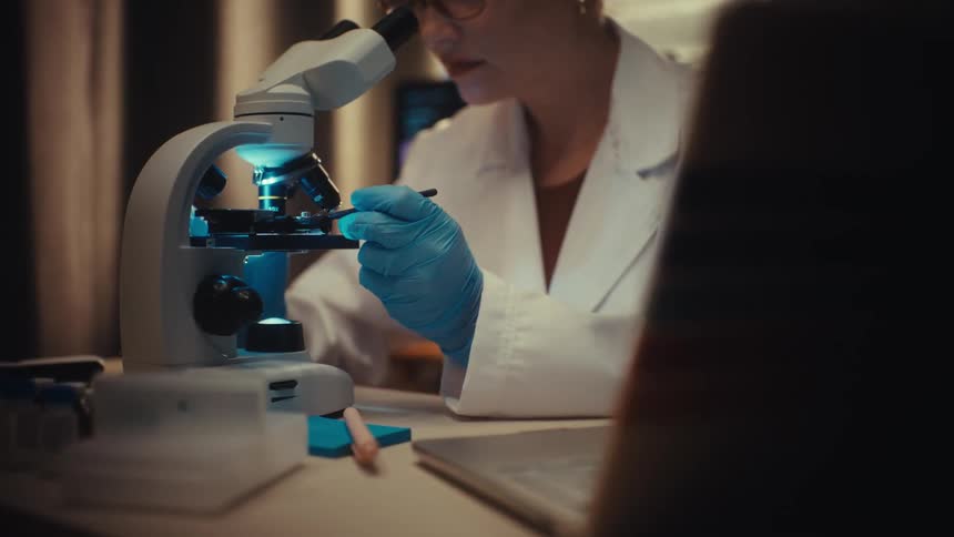 A focused female scientist in a white coat and gloves meticulously prepares a sample for a microscope in a dark laboratory