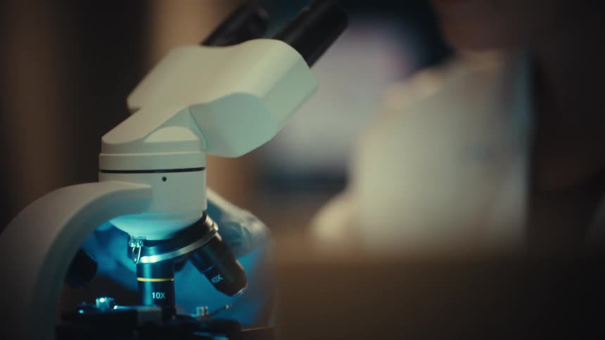 A close-up shot of a focused female scientist looking through a microscope in a dimly lit laboratory, with the camera racking focus between her face and the lens