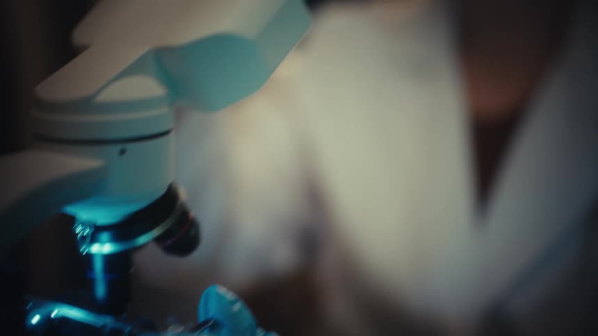 A scientist places a glass slide under a microscope and then holds it up to the light for close examination in a dimly lit lab