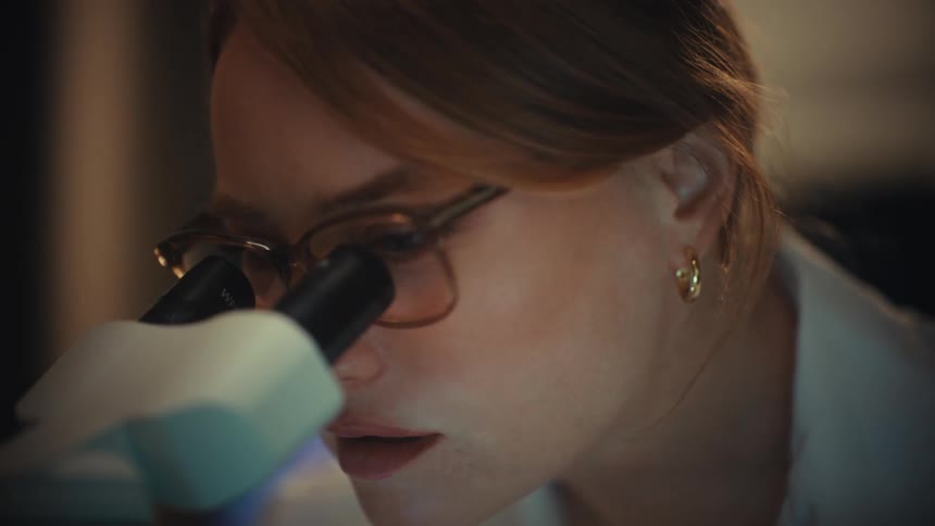 A female scientist in a lab coat looks into a microscope and smiles with joy at her discovery