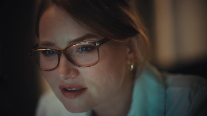 A dedicated female professional with glasses concentrates on her work on a glowing computer screen in a dark room